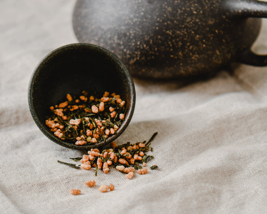 A steaming cup of Genmaicha tea in a traditional ceramic cup, with visible roasted brown rice and green tea leaves, capturing the rich, comforting flavor profile of this beloved Japanese brown rice green tea.